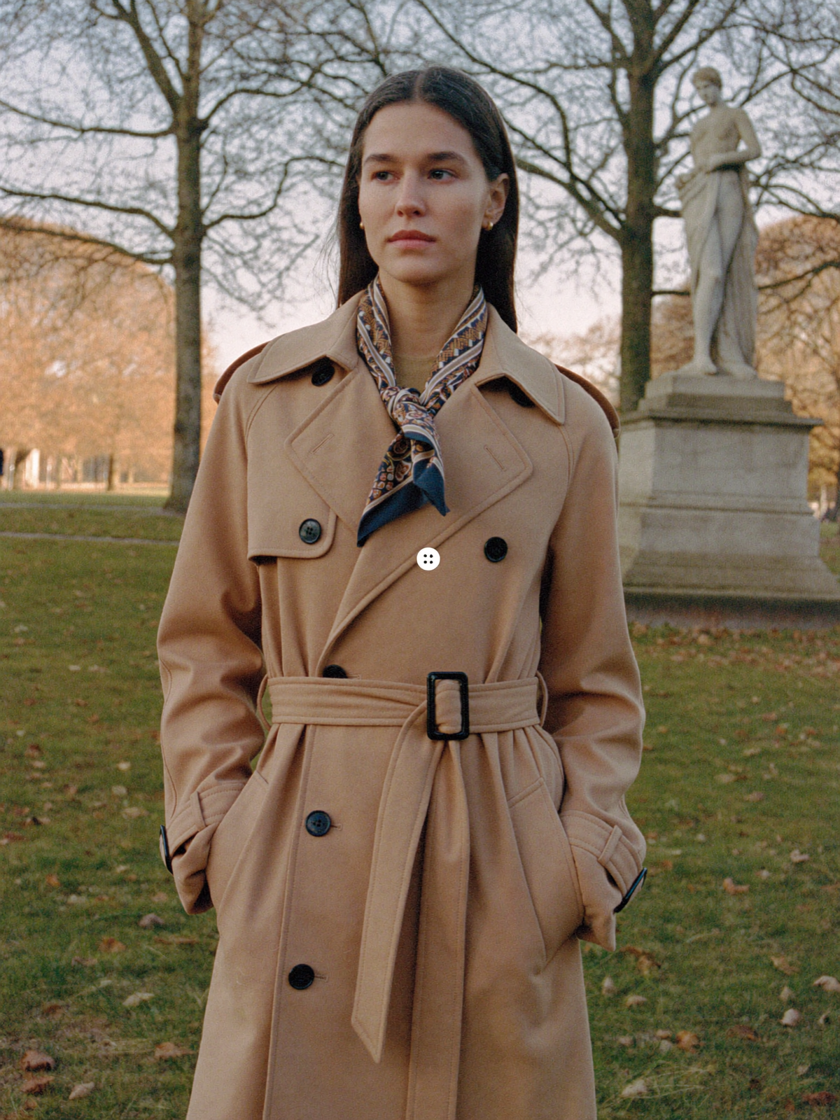 Woman in a camel trench coat with a silk scarf standing in an autumn park