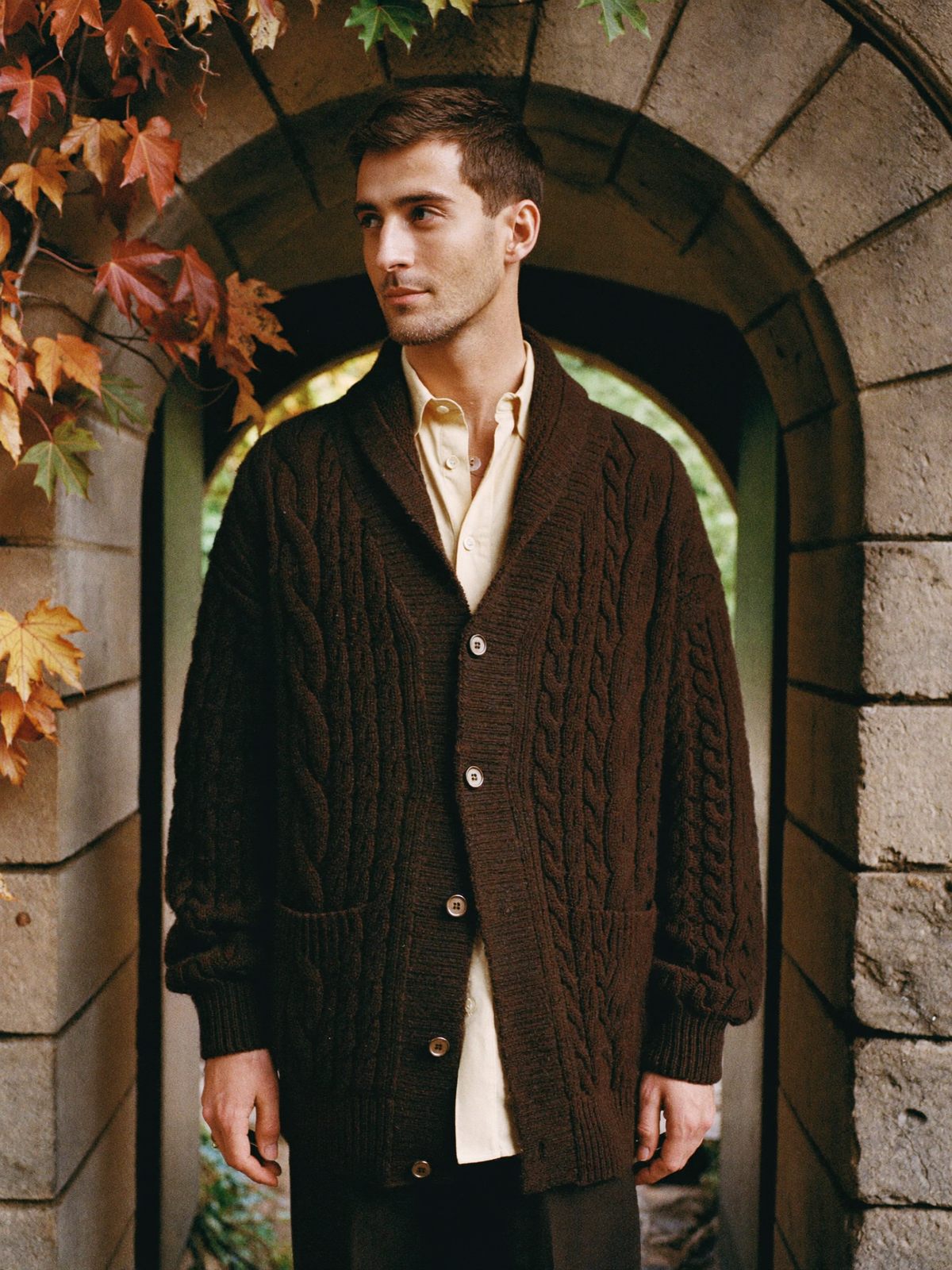 Man in a dark brown cable-knit shawl-collar cardigan standing under a stone archway with autumn leaves